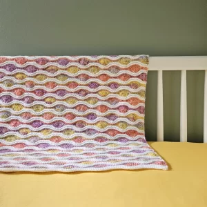 The wavy whimsy blanket draped on the side of a crib with yellow sheets and a sage green background
